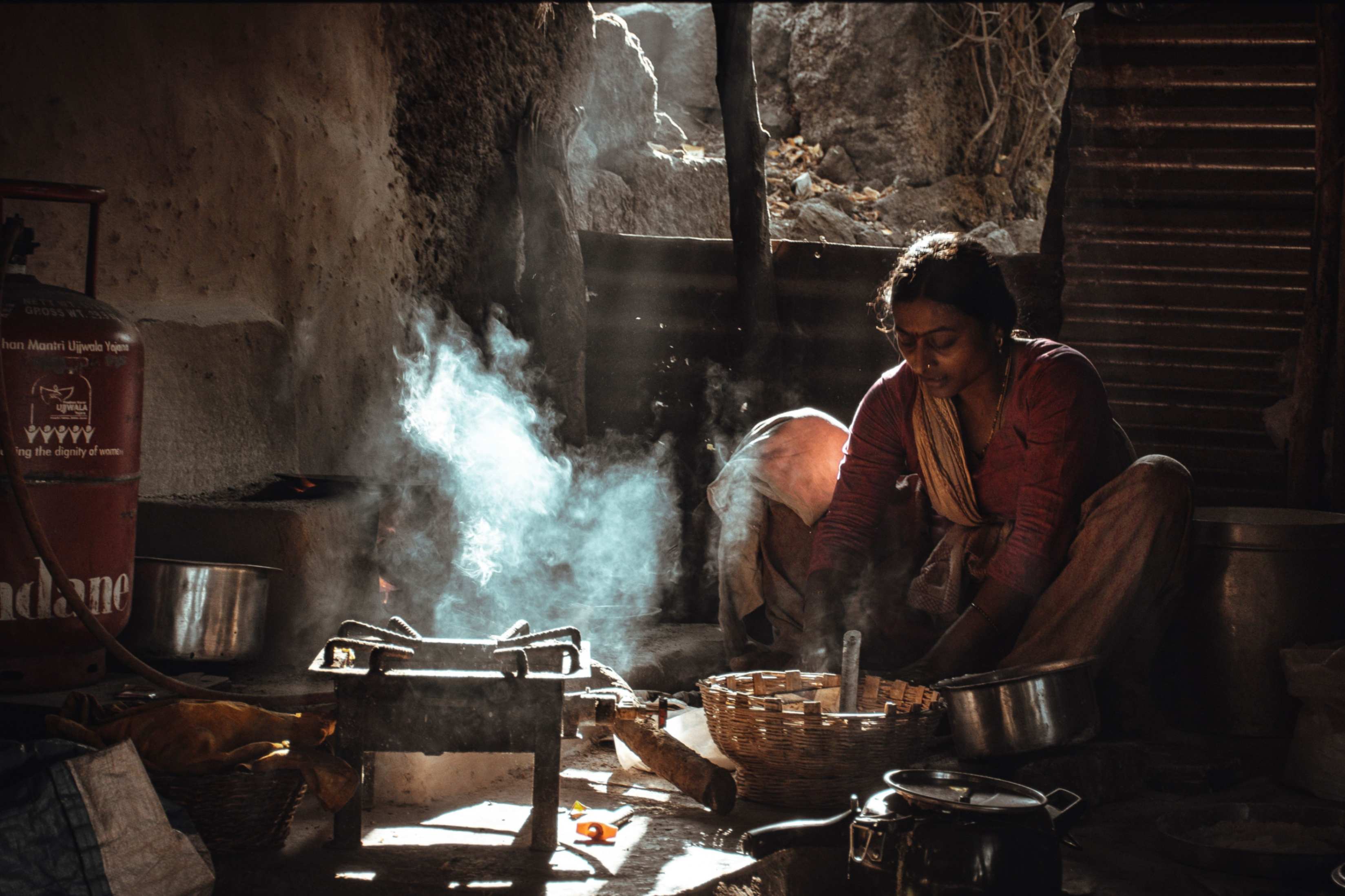 Una mujer cocina con leña en una vivienda rural de India, junto a un cilindro de gas del programa Ujjwala Yojana que permanece sin usar, reflejo de la escasez de GLP. (Foto: Pexels/Somya Dinkar)