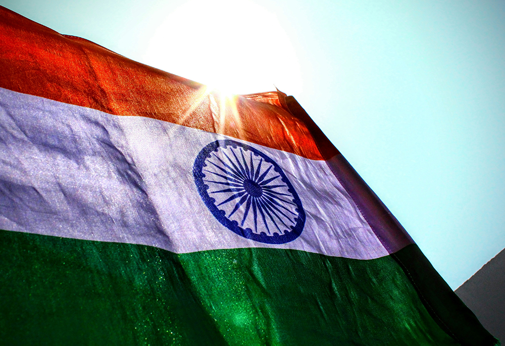 The Indian flag is backlit by the sun, with a blue sky in the background, in a close-up, outdoor photograph. (Unsplash/Aniyora J)