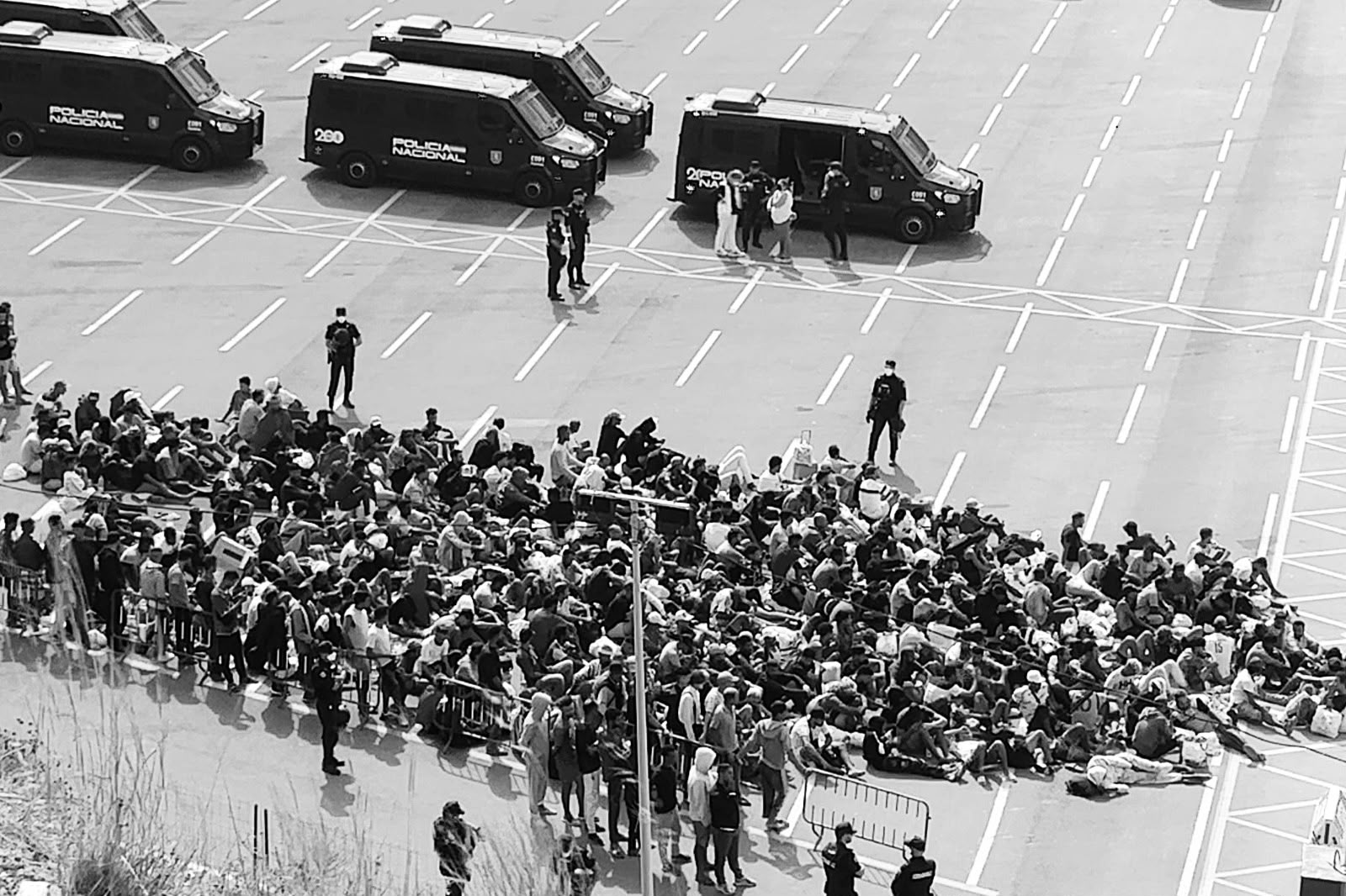 Decenas de migrantes permanecen sentados en el suelo de un estacionamiento, bajo la custodia de agentes de policía en Ceuta. El 11 de septiembre de 2026, el Gobierno de la ciudad informó de que unas 13 000 personas permanecían "en situación irregular o en recursos de acogida". (Foto: cortesía Mari Carmen Sapiña)