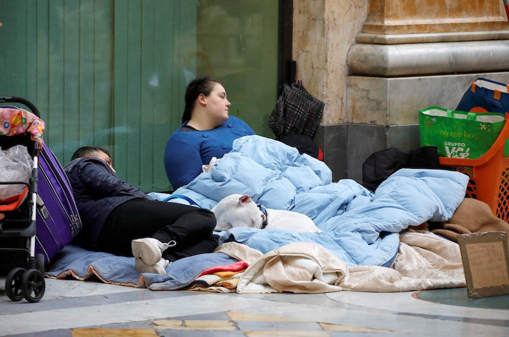 An adolescent boy lying on blanket on the street next to a reclining woman and bags of possessions