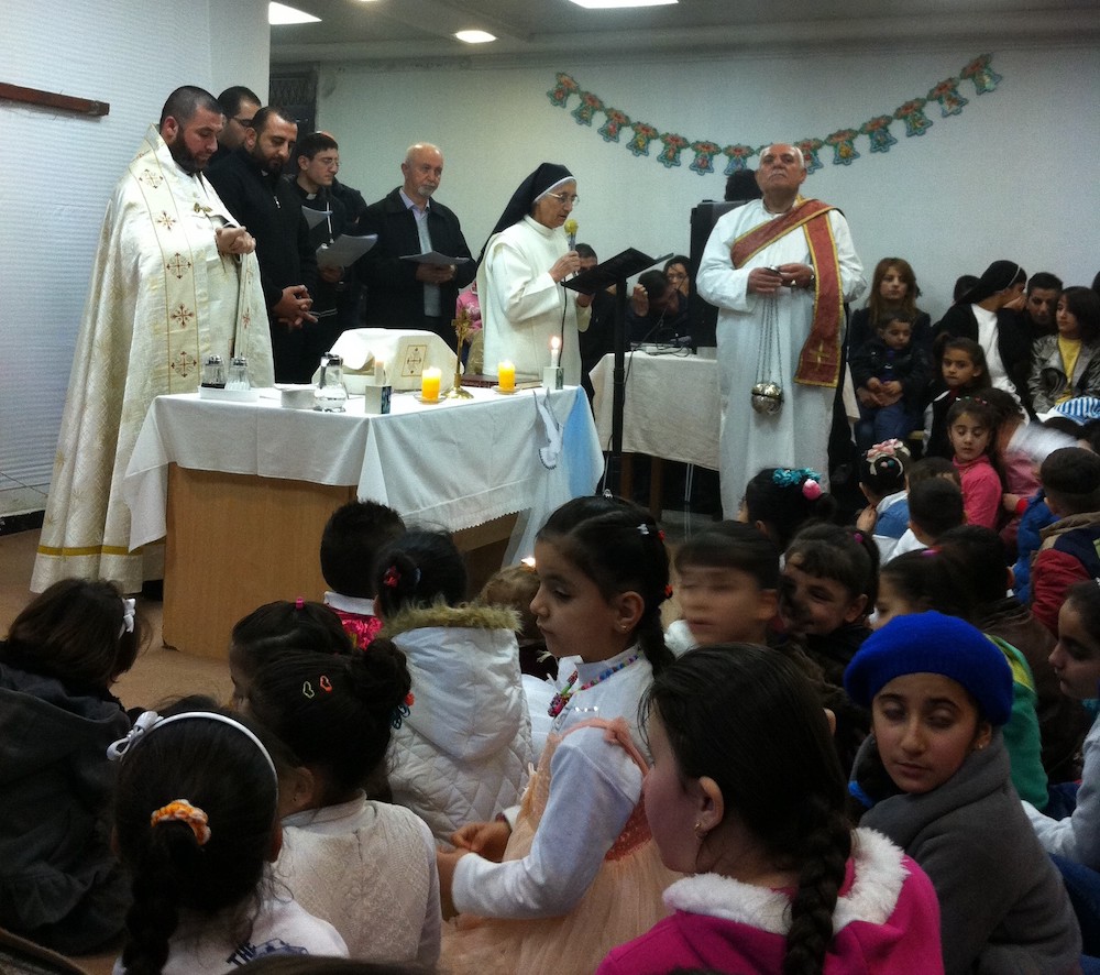 Priests at an altar while a nun in habit speaks into a microphone, with children sitting on the floor
