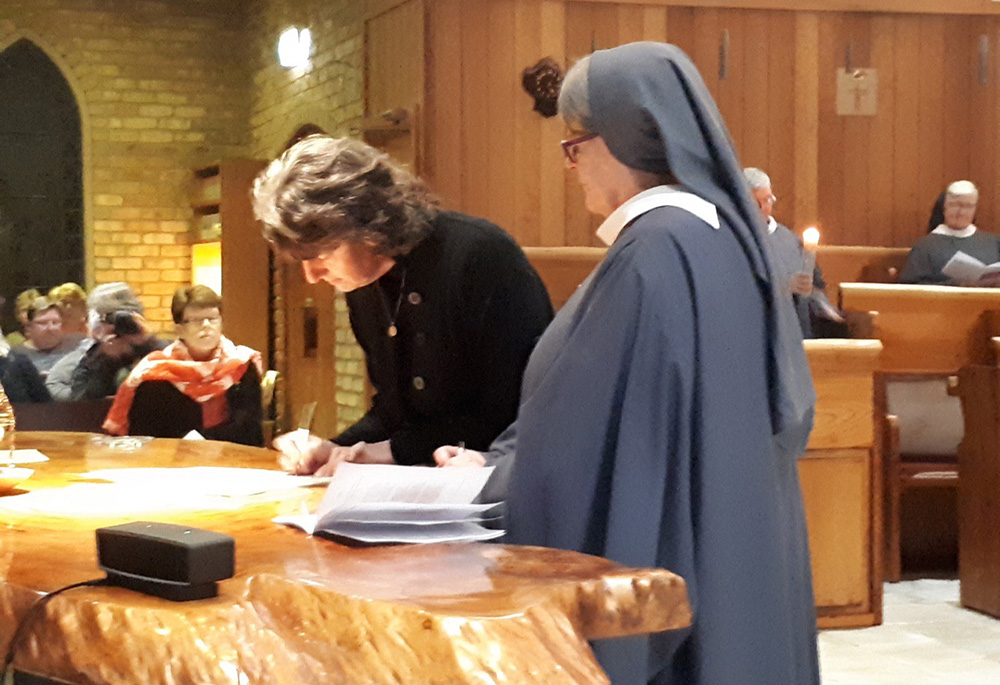 De pie y vestida de negro, Lee-Ann Wein makes firma un documento sobre un mesón, acompañada por una religiosa, al momento de hace sus promesas de beguina.
