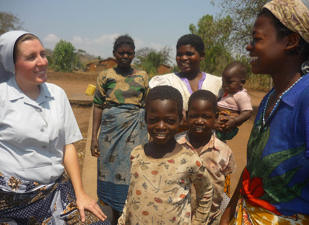 Sister Dwyer converses in an open field with young children and adults. She wears her habit and they wear colorful clothing.