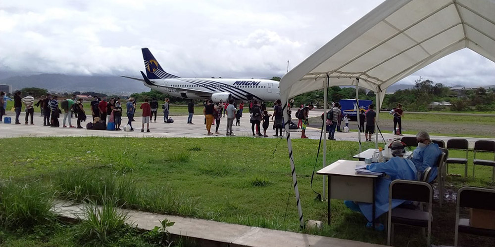 En una pista de aterrizaje, migrantes depoirtados hacen fila antes de subir al avión; a un lado de la pista, en un todlo blanco, la hermana Dondé y voluntarios scalabrinianos acompañan a los migrantes antes de abordar.