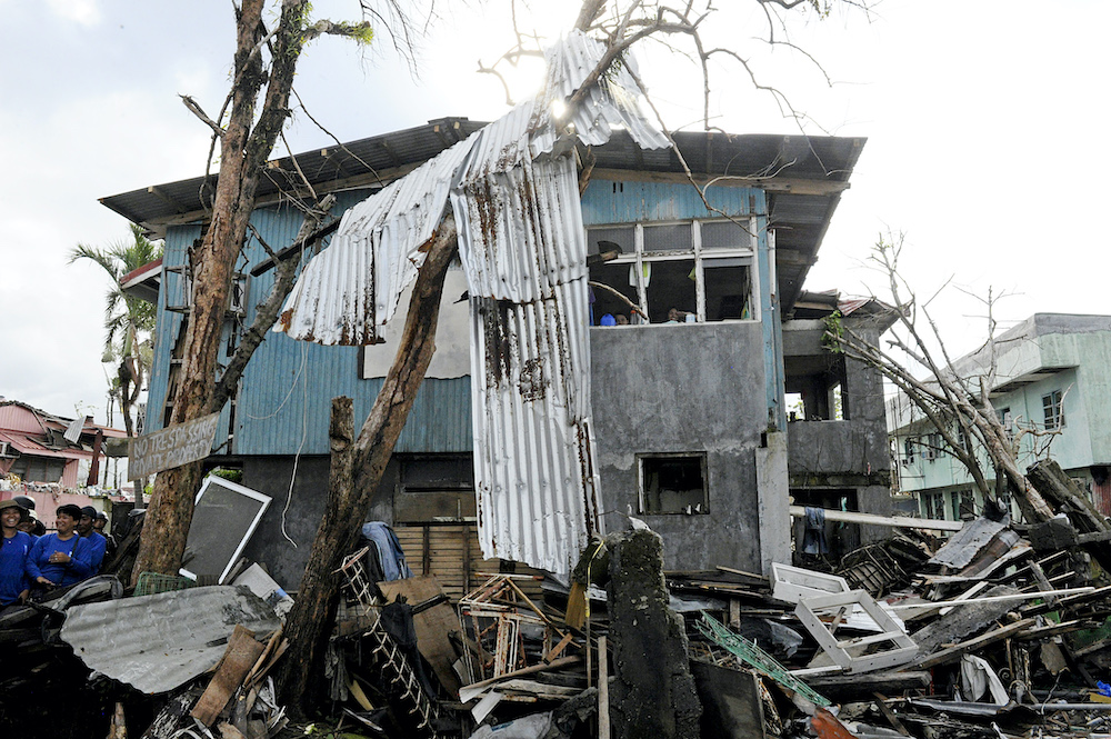 In this 2013 photo, residents of Tacloban peer out of a window of a damaged home as workers helping with clean-up efforts walk through the debris below following Super Typhoon Yolanda/Haiyan. On matters of housing and shelter, the Philippines is vulnerabl