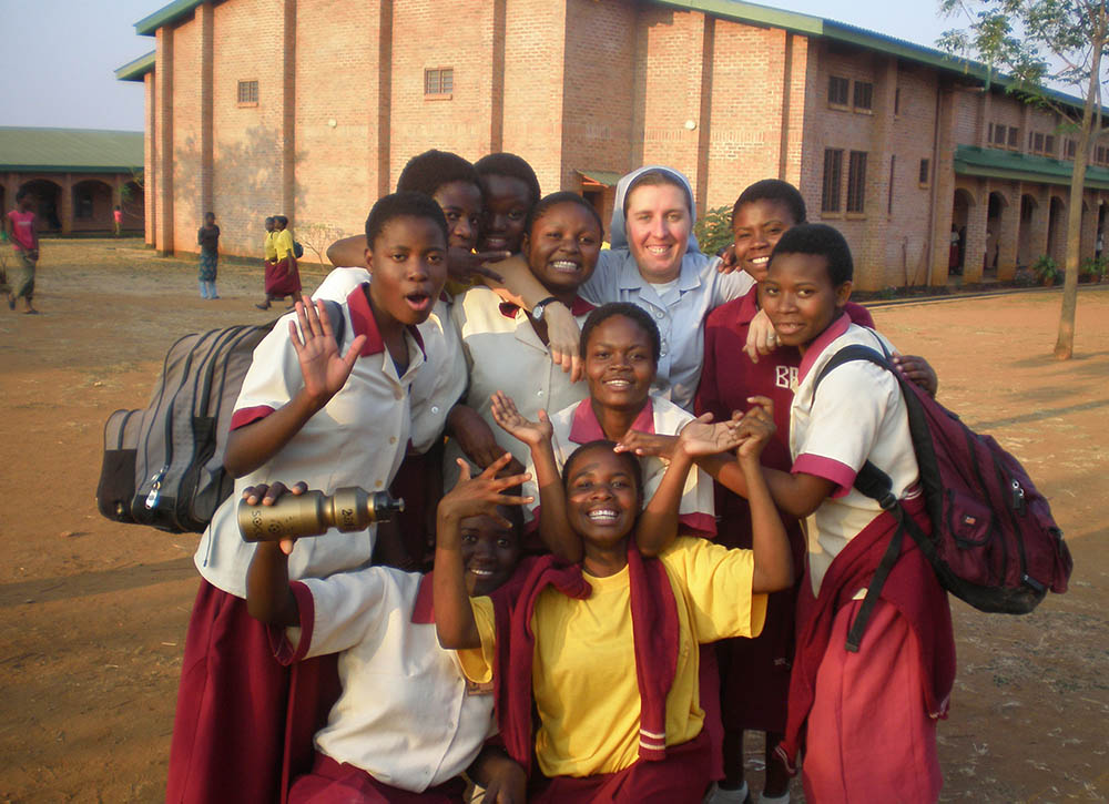 In the foreground Sister Dwyer dressed in her habit alongside students dressed in red and white; in the background their school. 