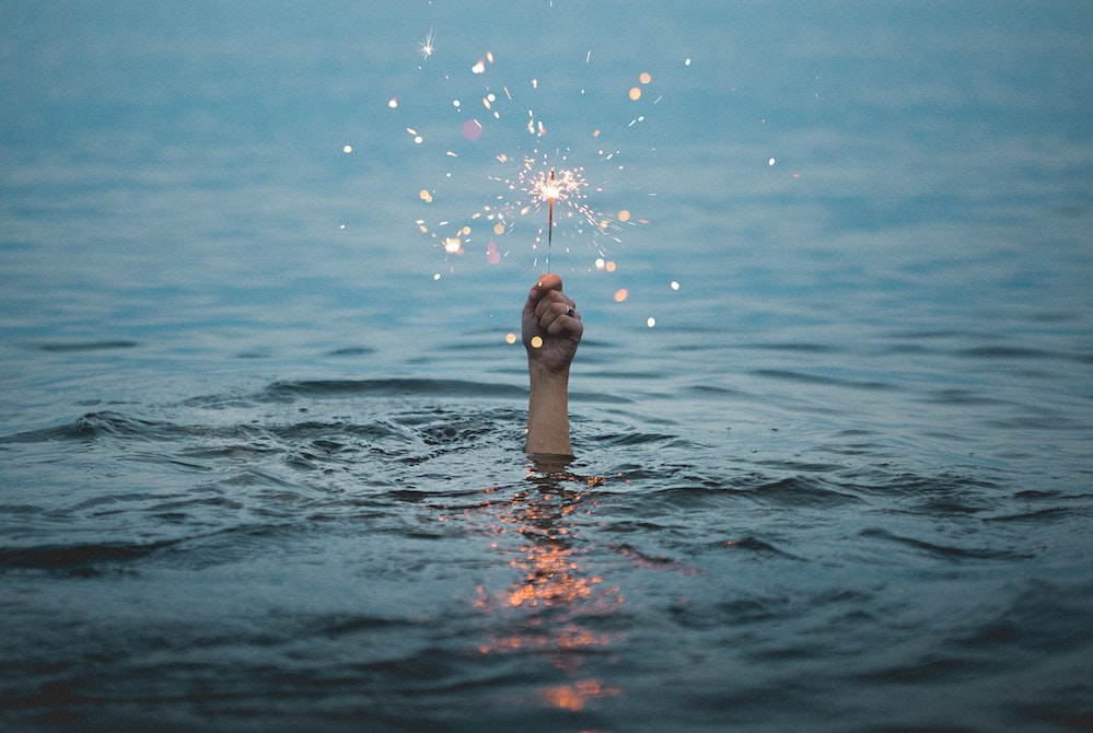 Image of a person holding a lit sparkler above the water (Unsplash/Kristopher Roller)