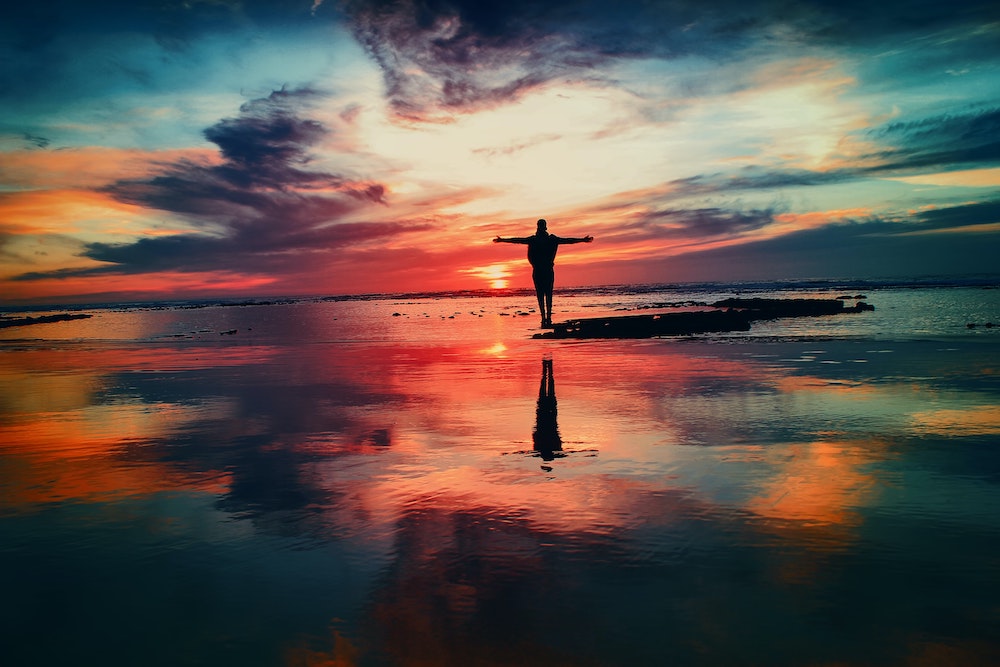 silhouette person on beach with arms outstretched amid a colorful sky