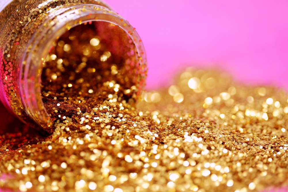 Image: A bottle of gold glitter lies on its side, glitter in a pile on a pink background.