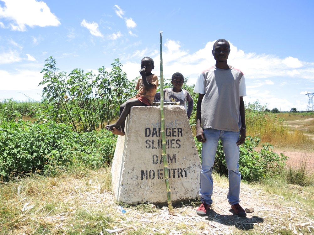 Children sit with some sugar cane on a sign warning against entry to the Lubengele Tailings Dam, next to the road leading from Chililabombwe to the border with the Democratic Republic of Congo. (Melanie Lidman)