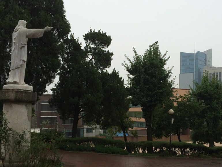Statue of the Sacred Heart stands in front of Sacred Heart High School across from tall building, headquarters for the Korean Racing Authority (Tom Fox)
