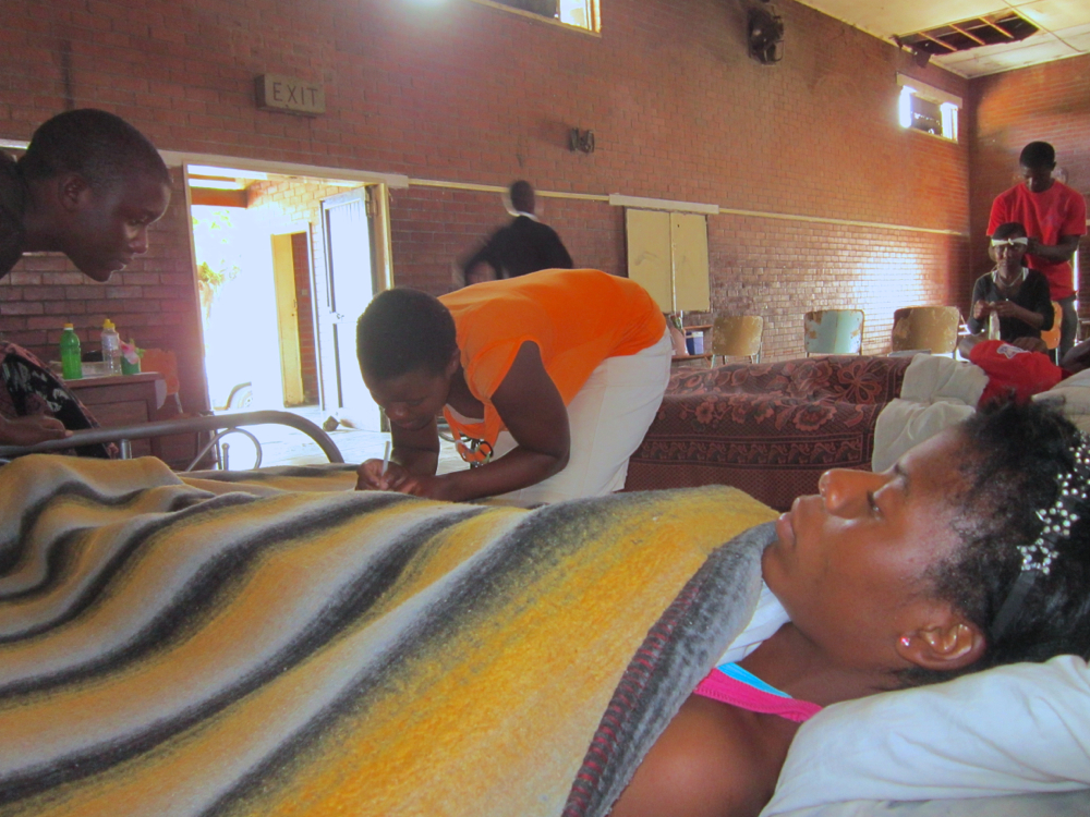 St. John trainers take everything they need to the community centers where they run Sr. Dominica Siegel’s home-based care courses for child-headed households. Here Kudzaishe Takaedza (left) and Lilian Maygadiena tuck in Fortune Washanyika while Rumbidzai Kamwendo practices bandaging Rutendo Rabaya’s head wound at the dilapidated community center at St. Mary’s Chitungwiza 12.4 miles (20 km) outside Harare. (Jill Day)
