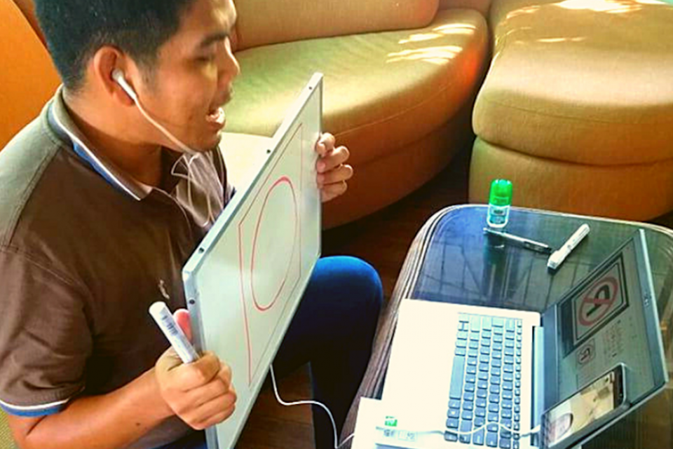 A young man at a laptop holds up a white board to teach an online class