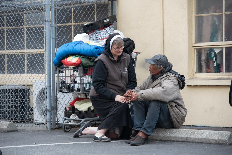 A habited nun kneeling to speak to an older man sitting on a curb next to a cart of his belongings