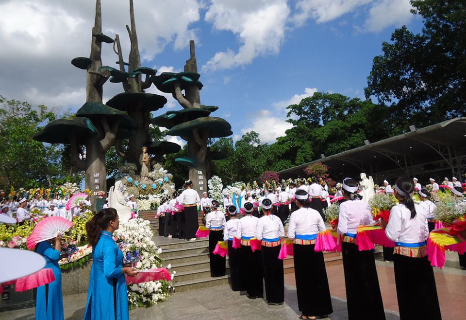 Women in traditional clothes dance with fans and flowers in front of a big statue of Our Lady of La Vang. (Joachim Pham)