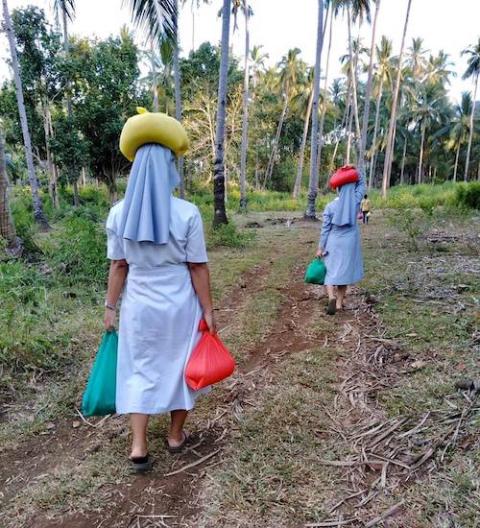 Two Catholic sisters in light blue habits walking on a dirt road carrying bags of rice