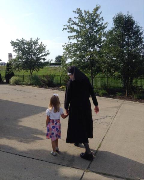 A nun in a black habit holds the hand of a preschool girl in a white top and floral skirt as they walk away from the camera