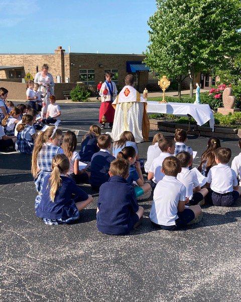 A group of school children in uniforms sit with their backs to the camera. A priest celebrates Mass in front of them. They are outdoors.