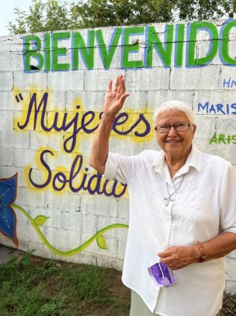 La hermana Virginia Bahena, vestida de nlanco, levanta la mano hasta su cabeza; detrás de ella, un mural con textos que dan la bienvenida a las mujeres solidarias. 