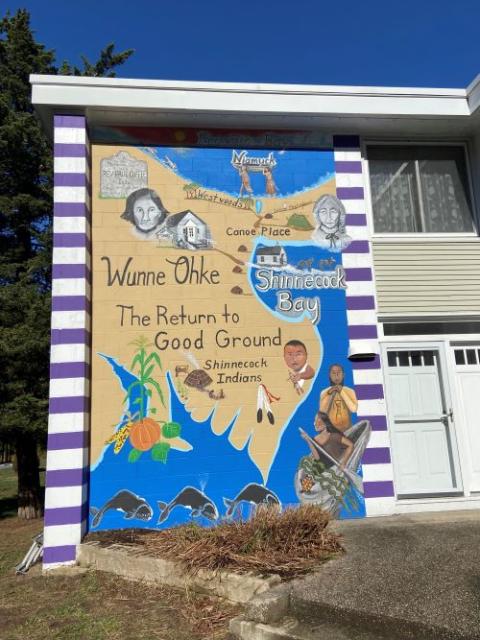 Mural called "The Return to the Good Ground" stands in front of a building. The mural looks like a map, showing land and water. 