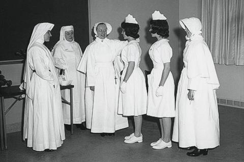 Nuns in white habits talk to women in white nursing uniforms in a black and white photo
