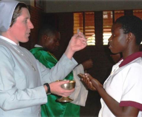 La hermana Dwyer en prmer plano, de perfil, con una hostia en su mano izquierda y un caliz en la derecha; frente a ella una joen estudiante y detrás un sacerdote. 