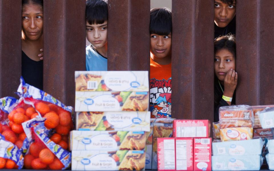 Brown people of different ages peer between the brown columns of the border wall at stacked boxes of food and bags of oranges.