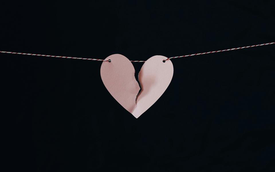 A pink paper heart hung on a string is pictured against a black background. The heart is torn nearly in two.