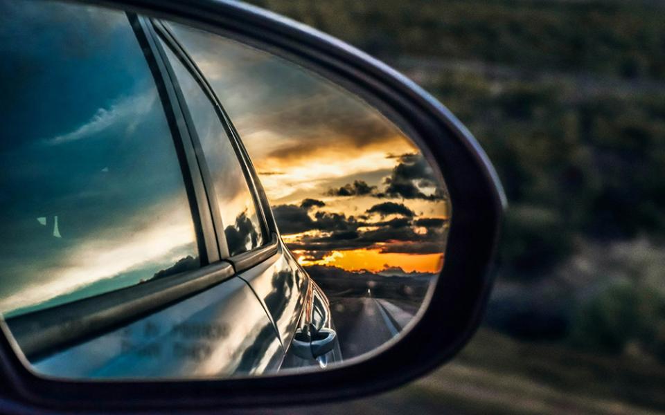 A car's rearview mirror shows a horizon full of bright orange and dark clouds. (Unsplash/Jake Weirick)