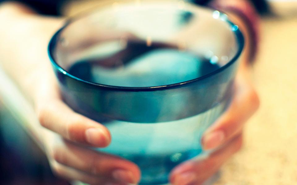 A photo presents an extreme close-up of a pair of hands holding and possibly offering forward a full glass of drinking water. (Unsplash/Amanda Yum)