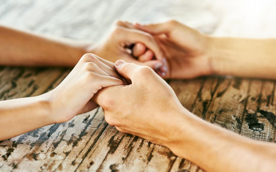 Closeup of two people holding hands across a wooden table (Dreamstime/Yuri  Arcurs)