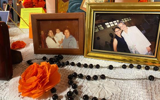 Two photographs sit on a table with a white lace tablecloth with a black rosary and orange poppy in front of them