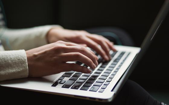 A Caucasian person wearing a white sweater types on a silver-and-black laptop keyboard. Only the hands are visible. 