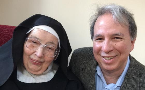 An elderly white woman in a black nun's habit and glasses poses with an older white man in a light blue shirt and gray suit jacket
