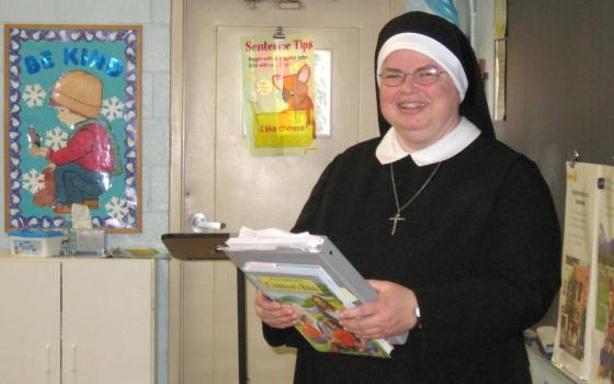 A white woman in a black nun's habit and glasses smiles at the camera, holding a gray binder and a book with both hands