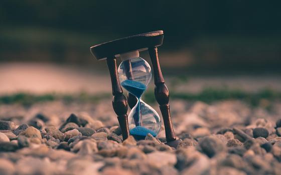 An hourglass with blue sand sits upon a field of rocks. The hourglass is half-full.