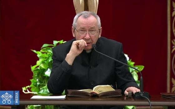 A priest in a black cassock speaks into a microphone in front of a burgundy backdrop