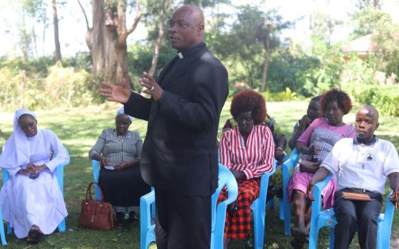 Priest stands and talks to a group of people sitting in blue chairs.