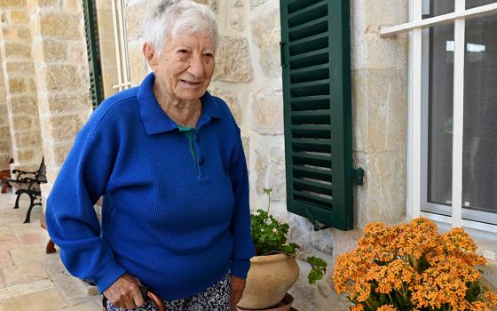 An elderly white woman in a blue long-sleeved shirt poses for a photograph next to a home with orange flowers