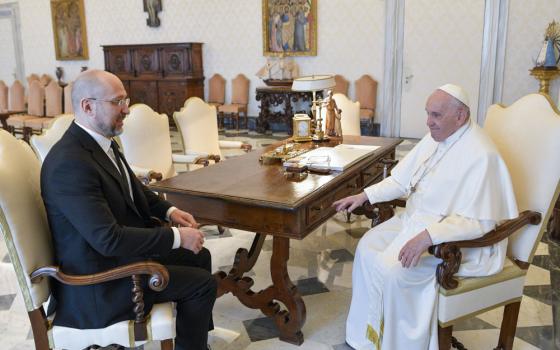 A white, bald man in a suit sits across from Pope Francis at the corner of a table