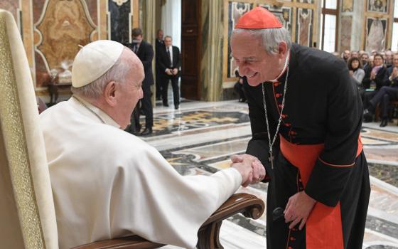 A cardinal leans down to greet a seated Pope Francis in a Vatican reception hall