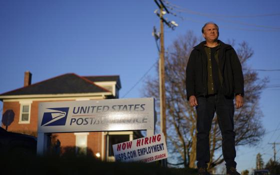 A white man wearing street clothes stands outside a United States Postal Office