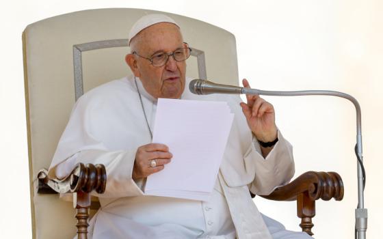Pope Francis sits in a tall-backed white chair, holds a paper, speaks into a microphone and makes a pointing gesture
