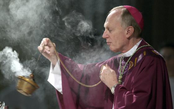 A white man wears a violet zucchetto and purple vestments and swings a thurible