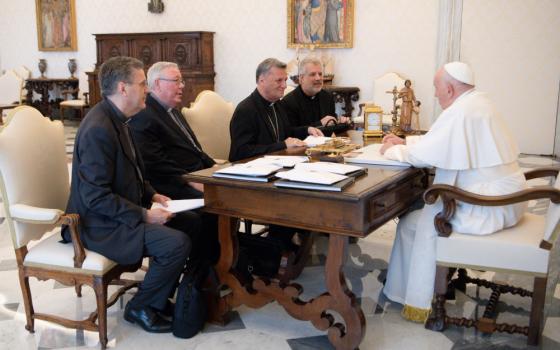 Pope Francis sits at his desk opposite four light-skinned men wearing black clerical garb