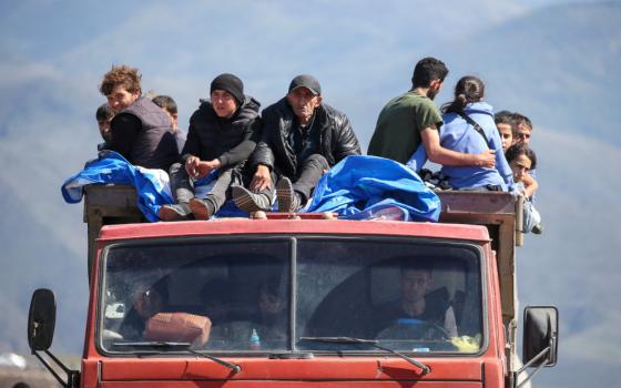 At least ten people sit on the roof of a orangey-red truck. More people are visible in the cab of the truck.