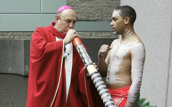 A white man wearing a pink zucchetto and red vestments blows into a digeridoo while standing next to a dark-skinned youth wearing white body paint.