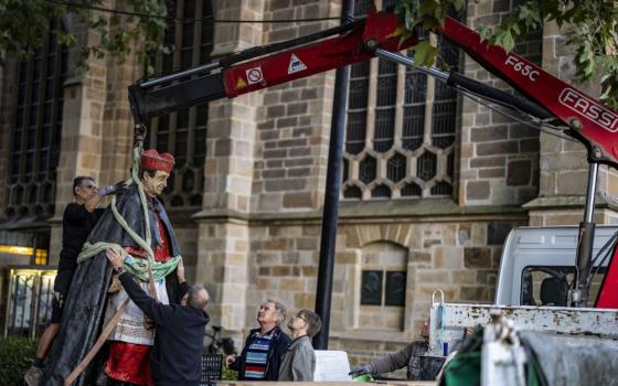 A statue of a man wearing a red zucchetto is lifted by a red crane while men steady the statue and others look on