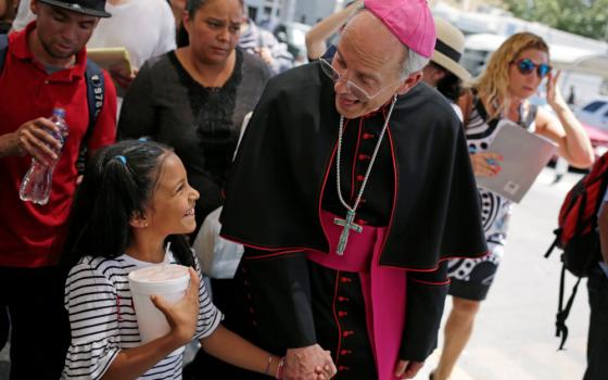 A white bishop wearing a house cassock looks down at a brown girl and holds hands with her as they smile at each other