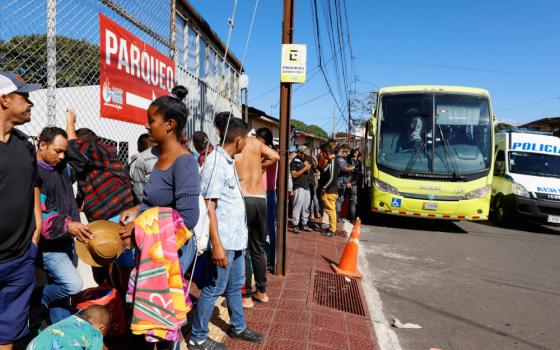 Brown people form a line on a sidewalk in between a chain link fence and a yellow bus